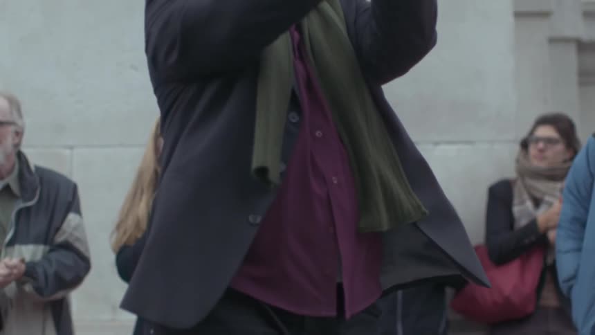 View of a man in a suit clapping his hands as others stand along a wall behind him and clap their hands