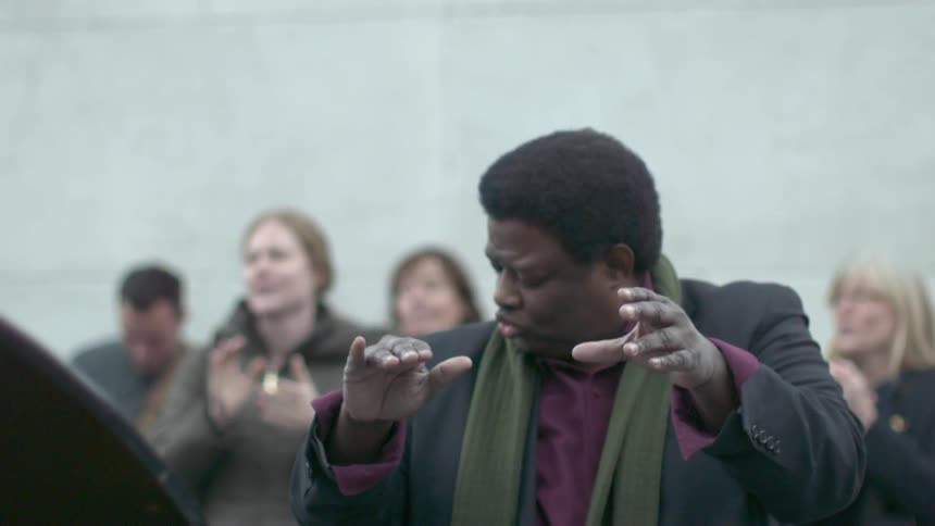 A man, dressed in a black suit and purple shirt, claps along with others while standing outdoors