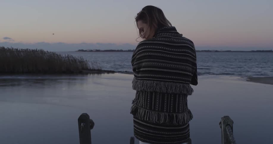 A young, blonde woman with a blanket wrapped around her looking over her shoulder and then downwards during sunset near the water