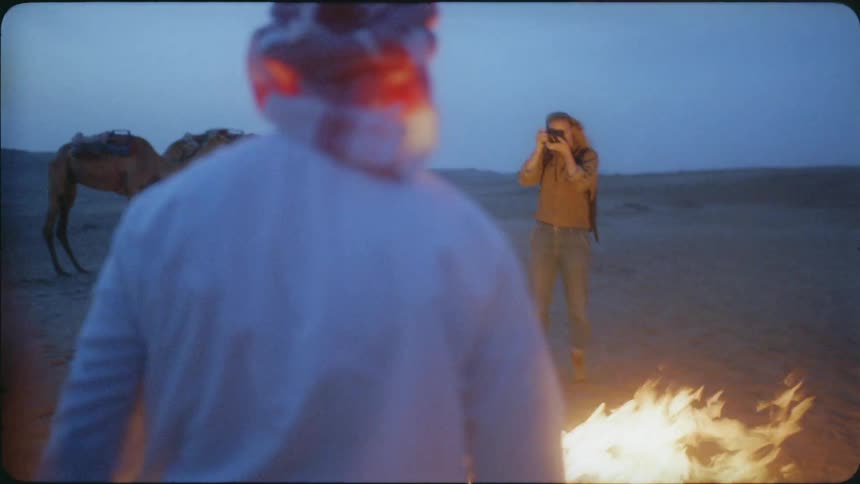 Woman stands outside in the desert near camels and takes a picture of a man in front of a fire