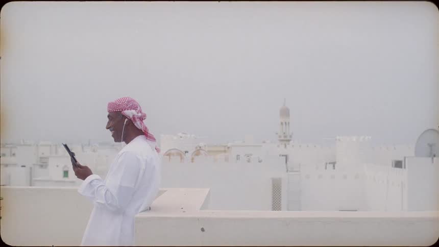 An arab leans against a rooftop wall and talks on his phone