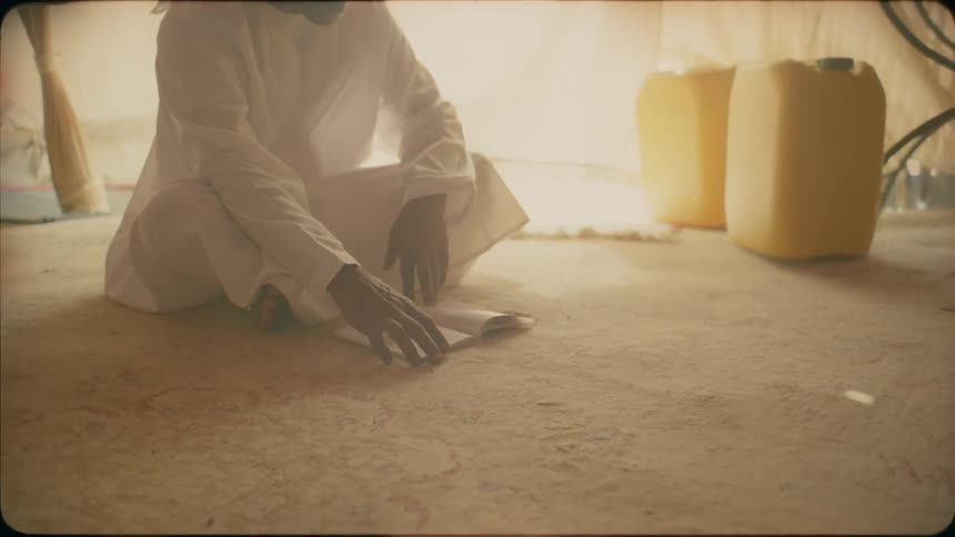 A man sits on a rug in a tent reading from a book
