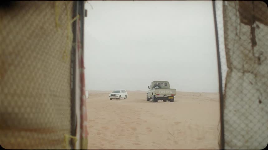 A woman sits by a tent near some vehicles in the sand