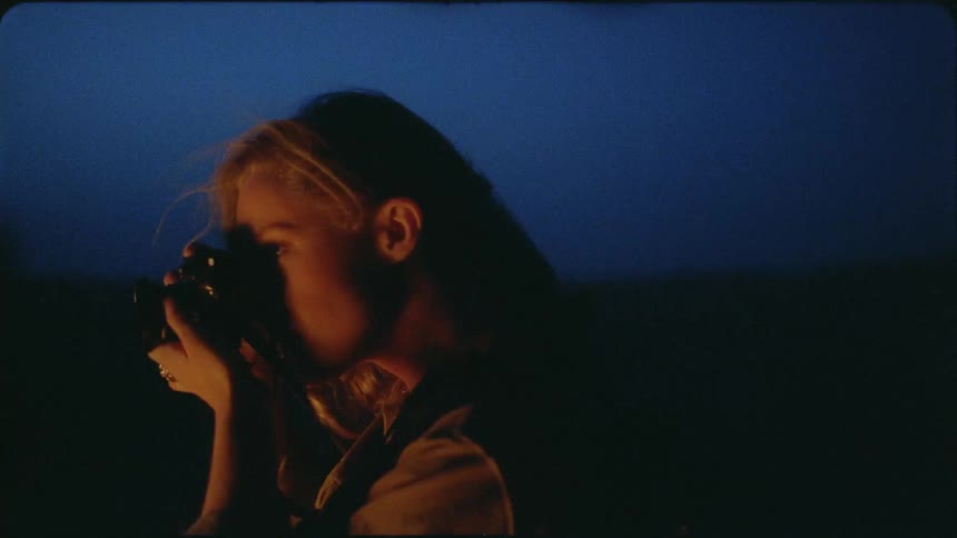 A woman focuses a camera and takes a photograph by a campfire at night