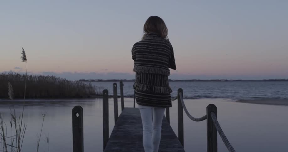 A woman, wrapped in a blanket, walking towards the end of a pier on a cloudy evening as the sun sets