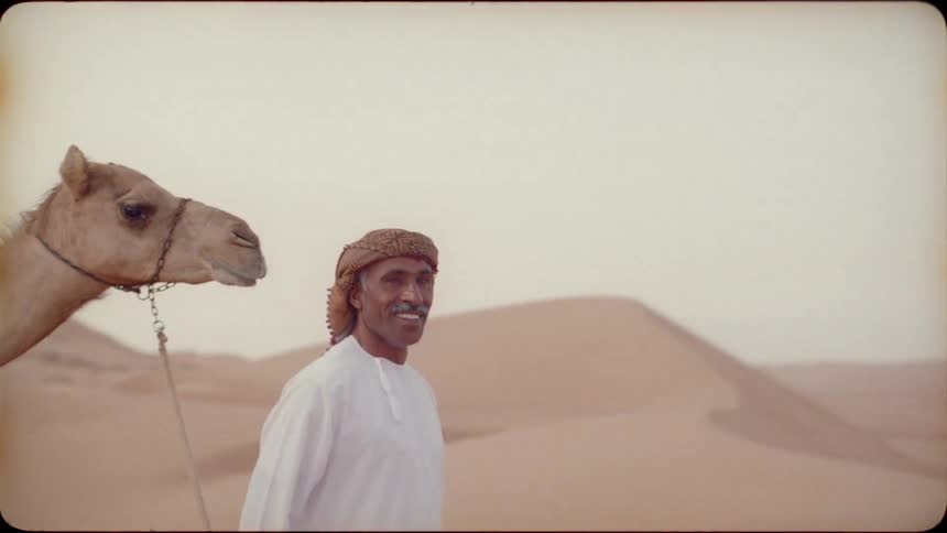 Man smiles and leads a camel through the desert as a woman watches