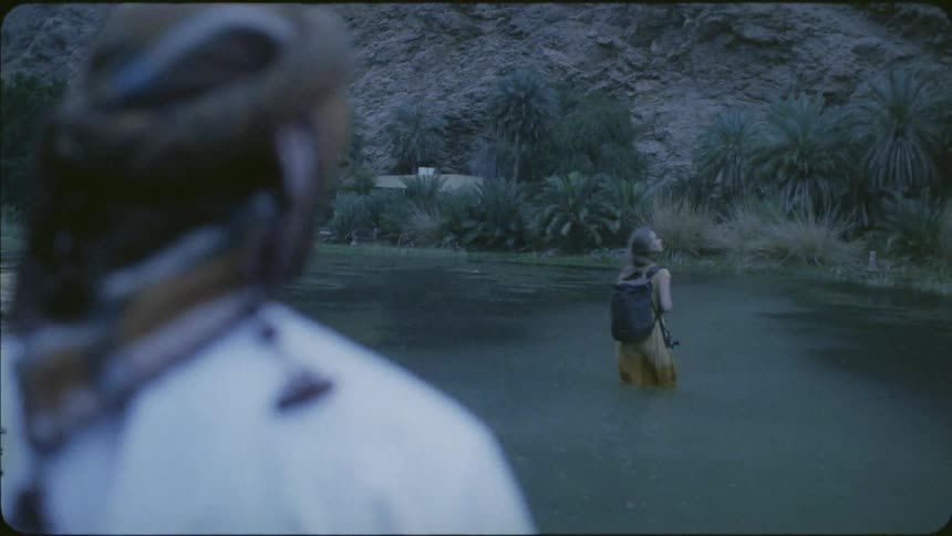 A man follows behind a woman wading across a river