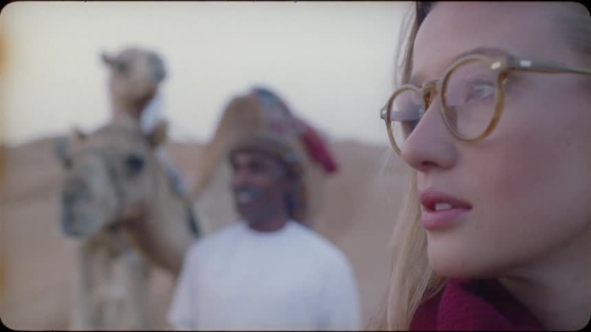 A woman walks in the desert with a man and camels