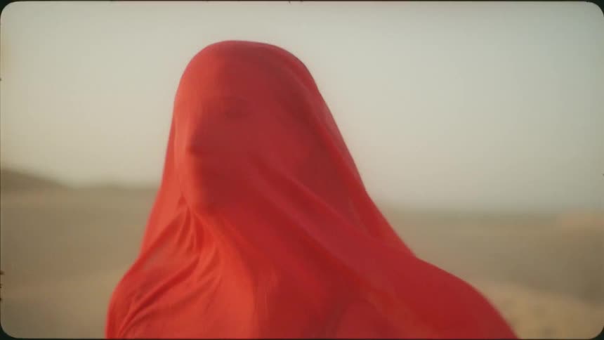 A woman stands outside in the desert with red fabric covering her face and blowing in the wind