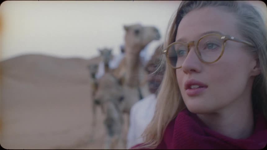 A woman walks with a man and camels in the desert