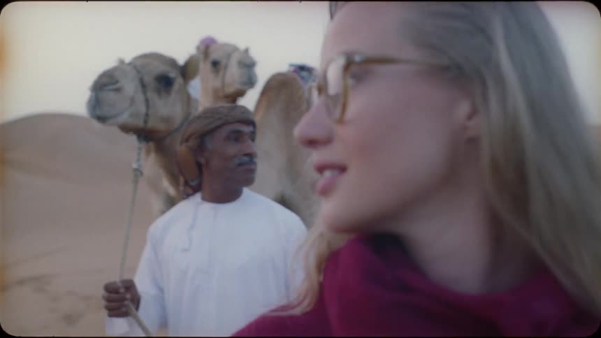 A man smiles at a woman as he leads camels through a desert