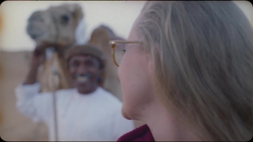 A woman laughs with a man who is standing near a camel in the desert