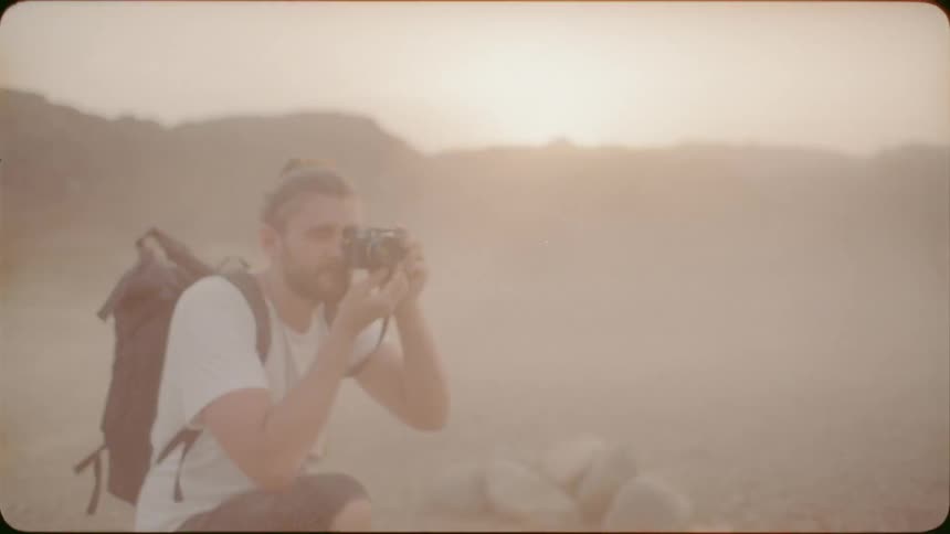 Man wearing a backpack kneels as he takes a photo with a camera