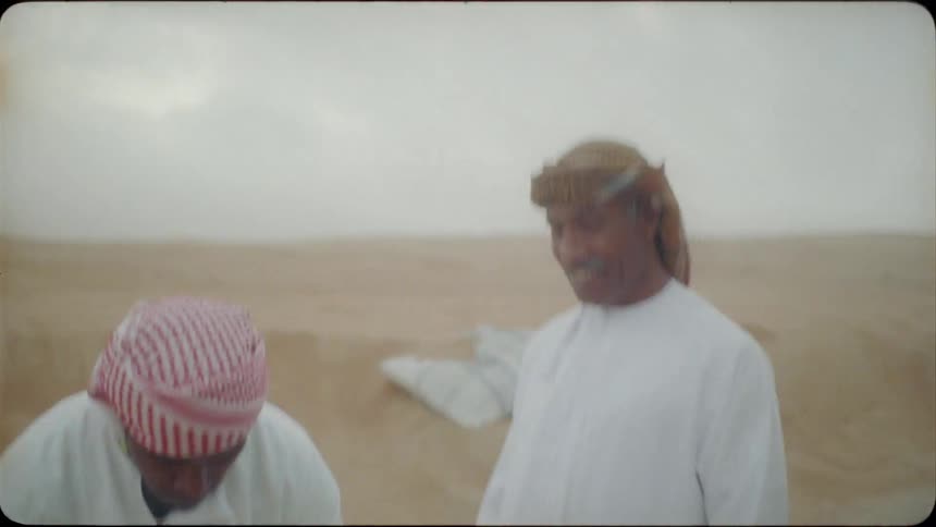 Two men kneel down in the sand to share a meal