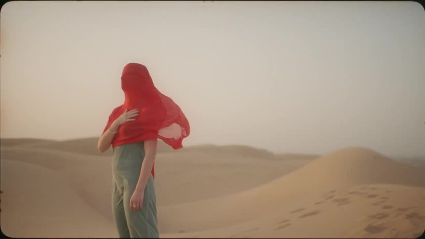 Woman stands still in the desert with a red cloth over her head