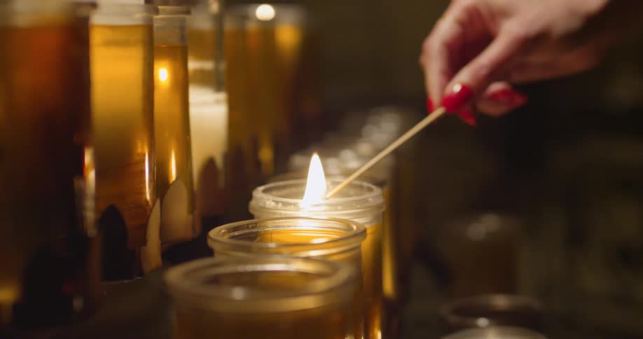 A woman with long, red fingernails lighting a candle that is in one of several rows of candles