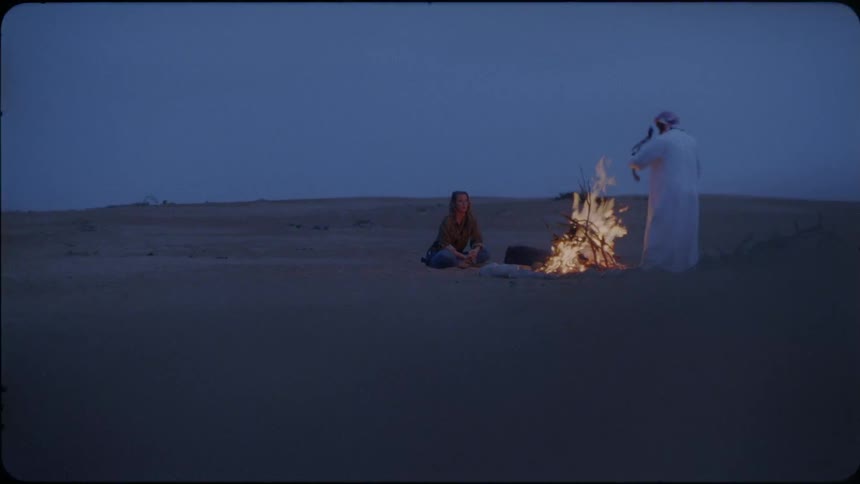 A woman sits near a campfire as a man puts branches into it