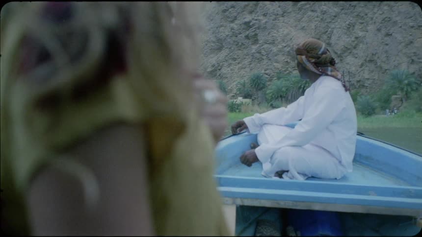 Woman stands outside near a man sitting on the roof of a vehicle by a lake