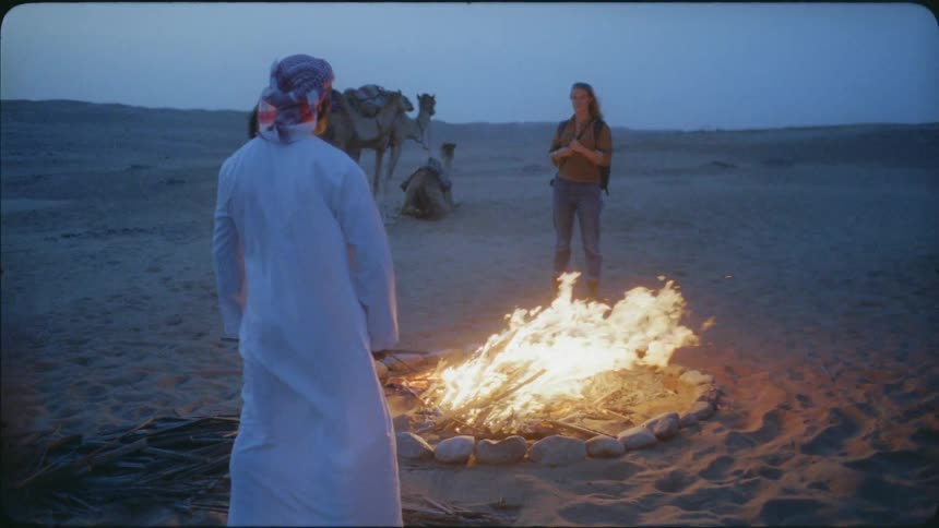 A man and woman stand by the campfire as camels rest