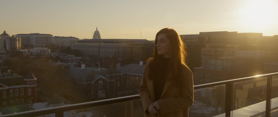 A woman is standing on a rooftop overlooking a city at sunset