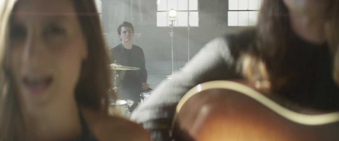 Man plays the drums as a woman sings while next to a man playing a guitar