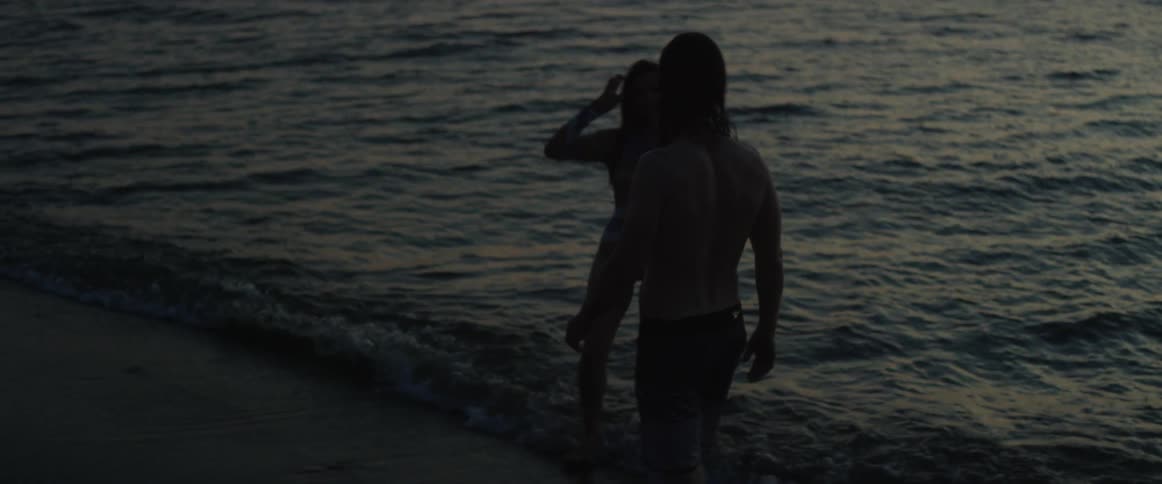 A man and woman are wading in water at the beach at dusk