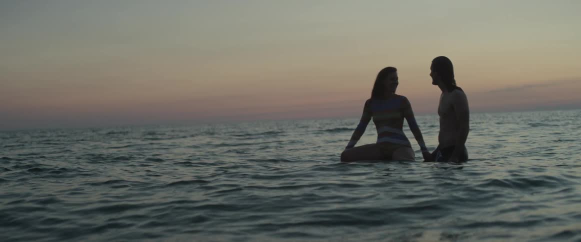 Woman sits on a surfboard in the ocean as a man stands next to her