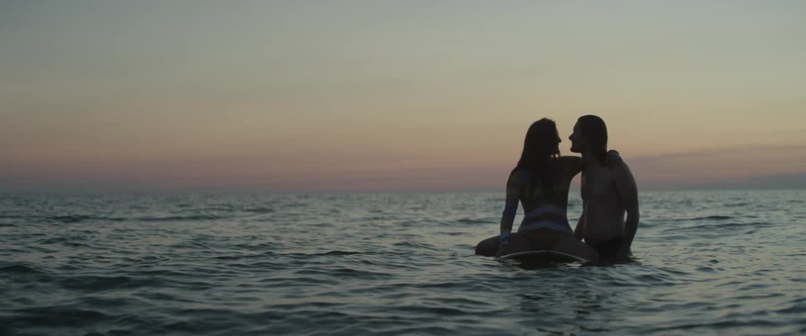 A man stands in the water beside a woman sitting on a surfboard holding hands
