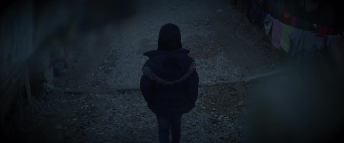 A boy is wearing a winter coat as he walks along a path inside a camp in daylight