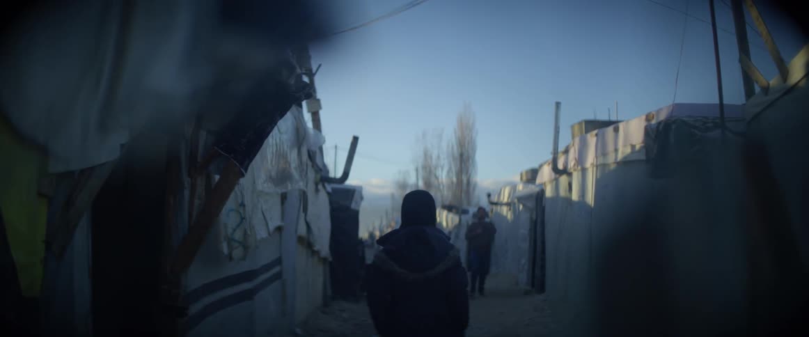 A person is wearing a coat as they walk through a refugee camp in daylight