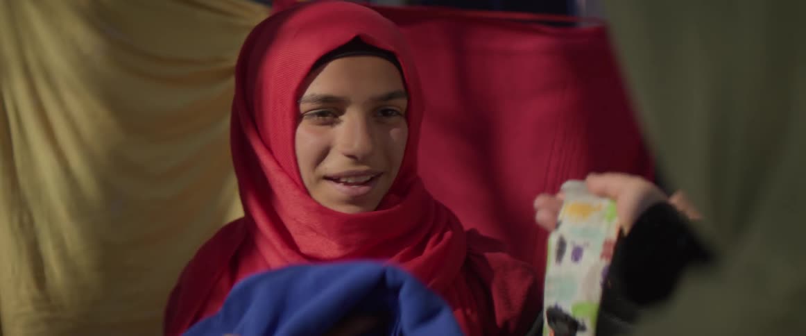 Child wearing a red headdress smiles and holds a blue cloth