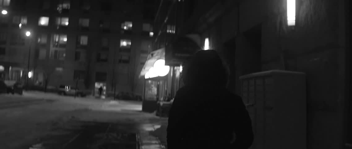 A person in hooded winter coat walks down a city sidewalk at night