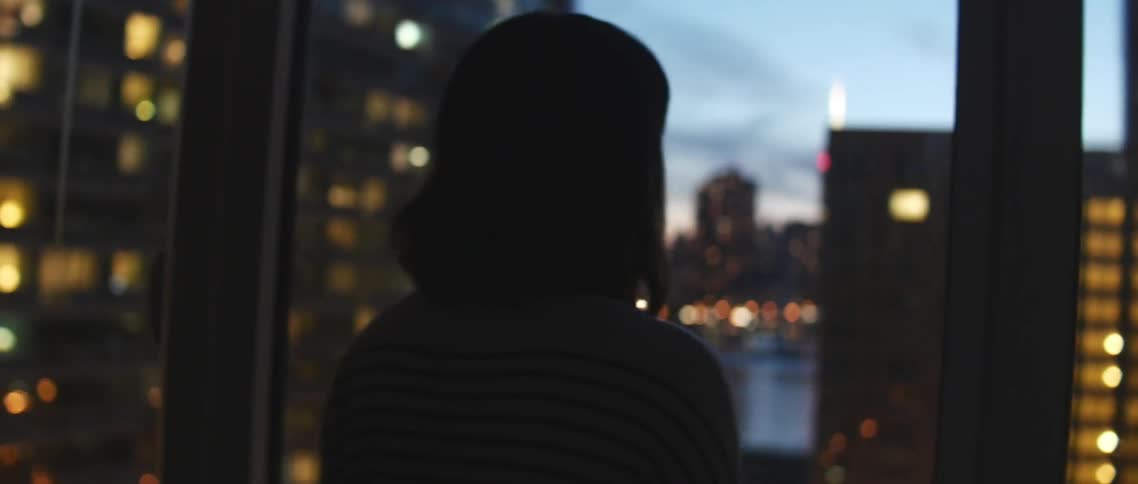 A woman walks toward the large windows and looks out at the city at night