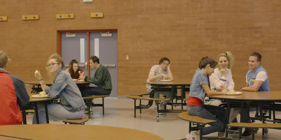 A boy sits alone at a table in the school cafeteria while others talk