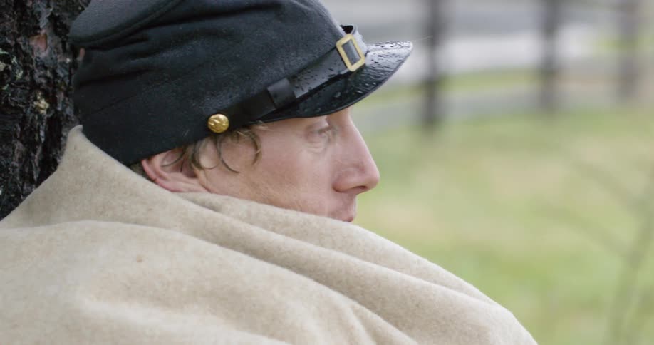 A man rests sitting against a tree with a blanket over him