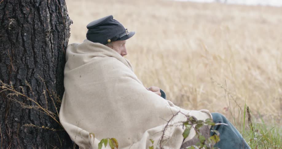 A man is sitting by a tree with a blanket over him