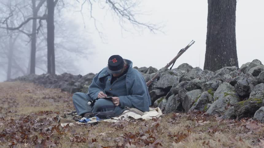 A soldier sits on the grass near a rock wall writing in a journal