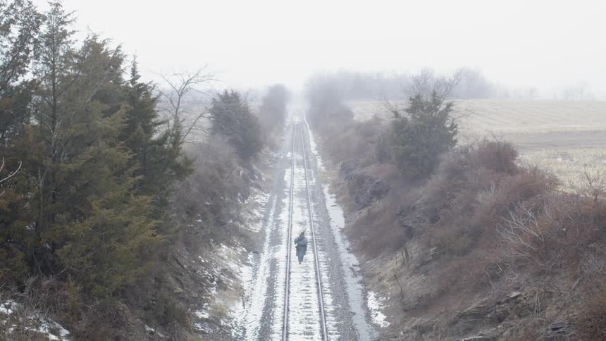 A person carrying a rifle walks down train tracks on a cloudy day