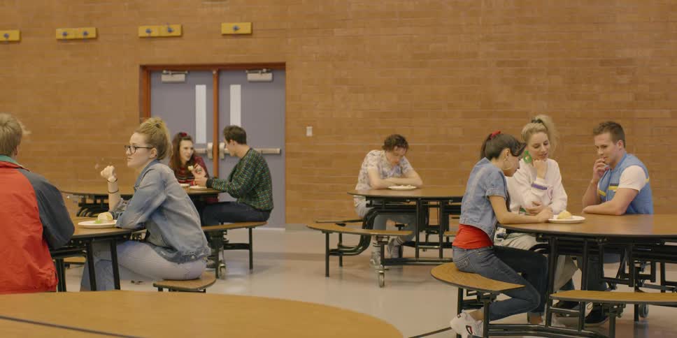 Teenagers sit at tables in a school cafeteria and one boy sits alone
