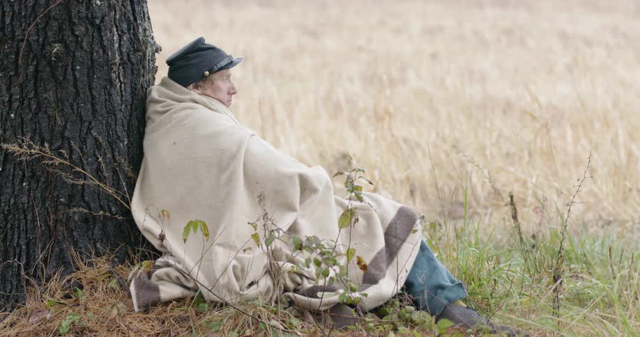 A civil war era soldier wrapped in a blanket leans against a tree