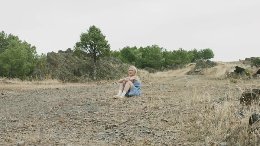 A girl sits in a clearing with trees and brush behind her