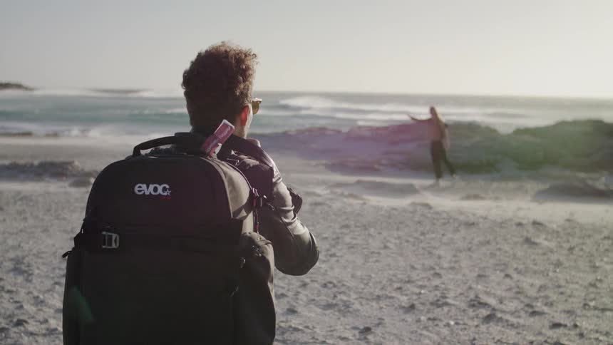 A man directs a woman standing on a beach and photographs her