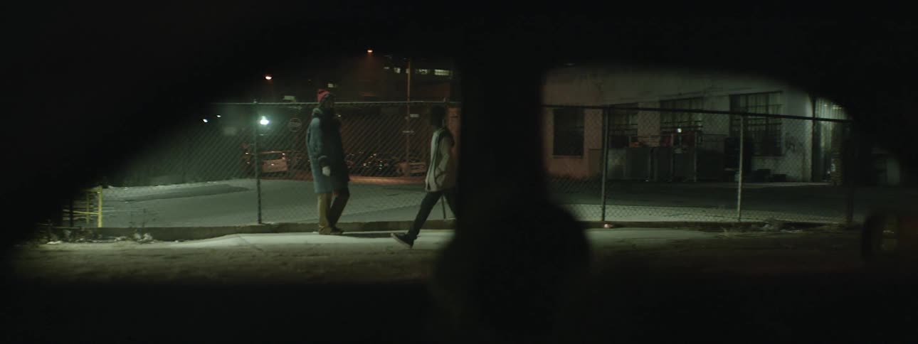 Man walking down a sidewalk past another person near a fence at night