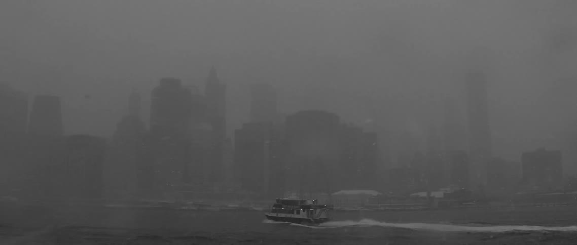 A boat cruising towards a coastal city on a very dark and snowy day