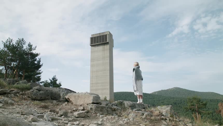 A woman stands on a rock near a cement tower and trees