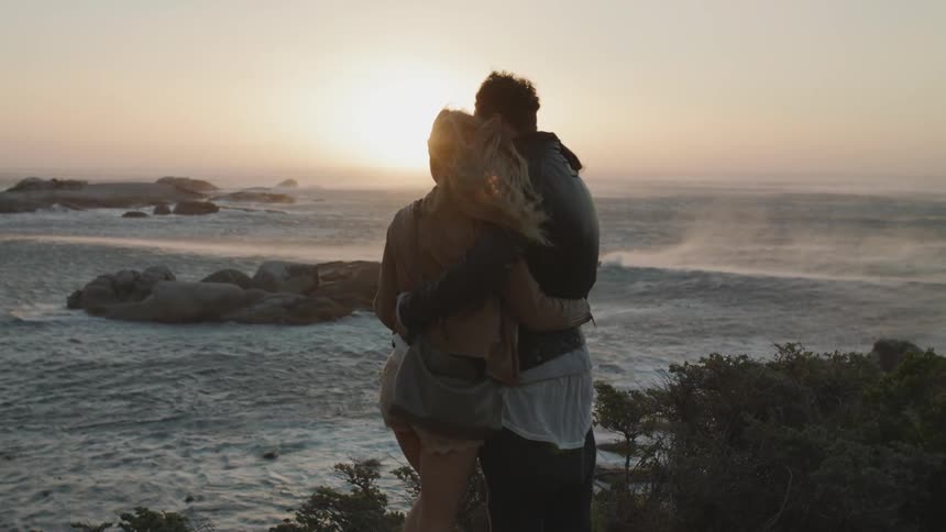 A couple a hugging each other as they stand watching a beach on a windy day