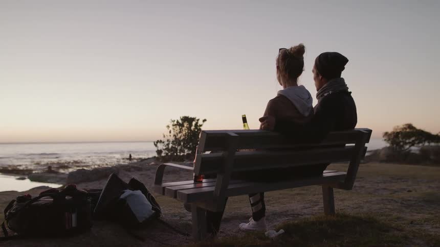 A couple are sitting on a bench drinking and watching the ocean waves at sunset