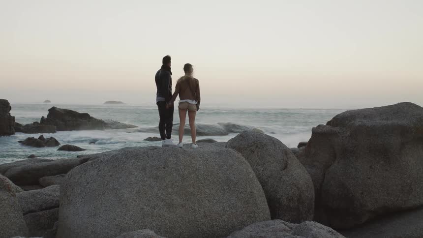 A couple hold hands atop a rock on the shore