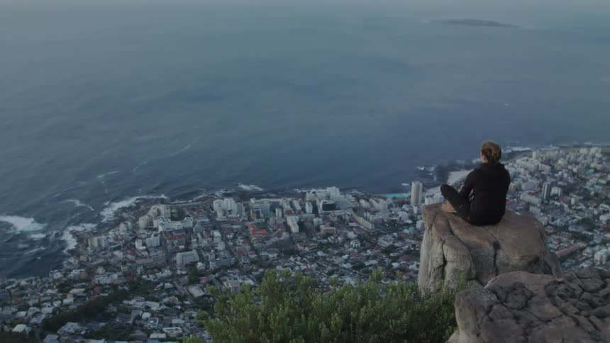 Woman sits on a cliff edge overlooking a coastal city
