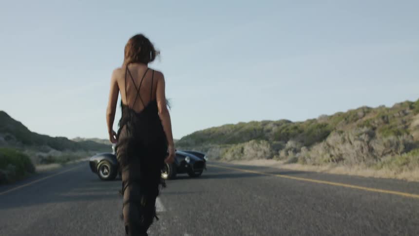 A woman in a fringed jumpsuit walks toward a man leaning against a convertible